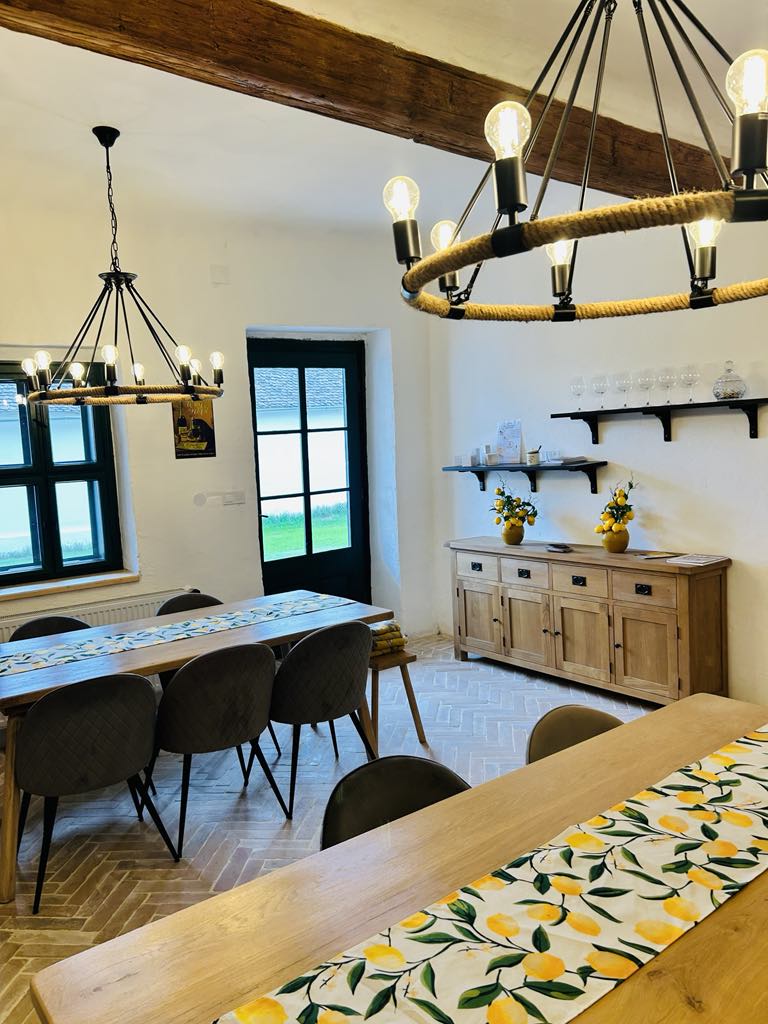 A spacious dining area features two long wooden tables accompanied by upholstered chairs. A large chandelier with exposed bulbs hangs overhead, while a sideboard displays decorative items and fresh flowers. Light streams in through a multi-pane window, illuminating the warm tones of the room.
