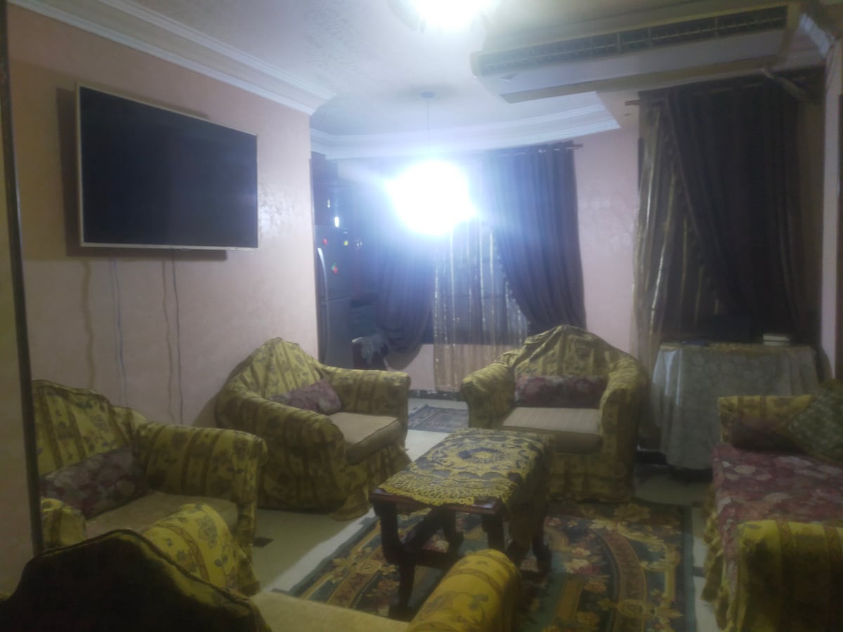 A living area features several patterned armchairs and a low table set on a decorative rug. A wall-mounted television is positioned above a side table. Natural light filters through curtains, while a ceiling fan adds to the comfortable atmosphere.