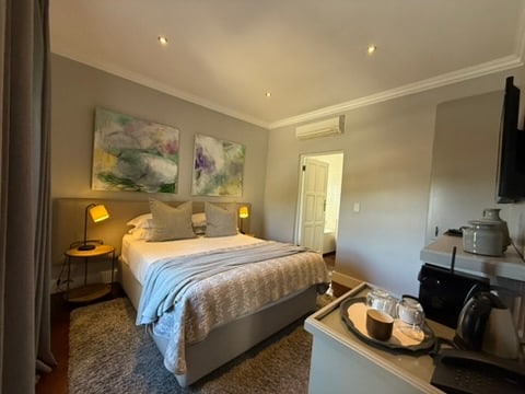 An intimate room is presented with soft lighting and muted grey walls. An XL Queen bed features plump pillows and a textured blanket. Two colorful artworks adorn the wall above. A small table holds tea and coffee-making facilities, complemented by glassware and a minibar.