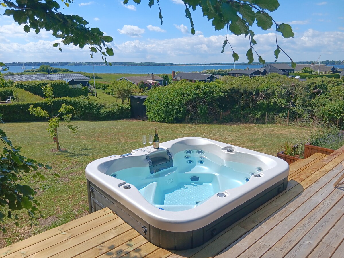 A spacious hot tub is situated on a wooden deck, overlooking a lush green lawn and the scenic Flensborg Fjord in the background. The area is framed by trees, adding a natural touch to the relaxing setting.