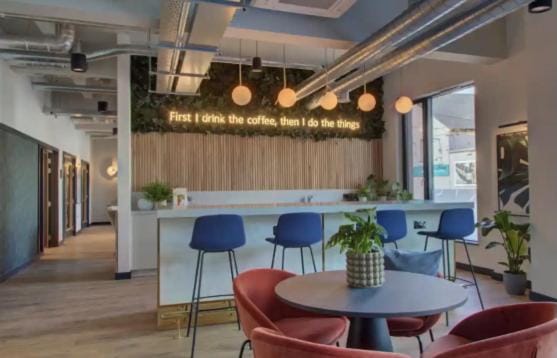 A communal lounge area is featured, highlighted by modern decor and warm lighting. The seating includes colorful chairs arranged around a circular table, while a bar counter provides a cozy space for refreshments. Greenery adds a natural touch to the environment.
