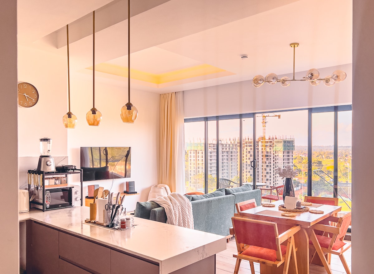 A modern kitchen integrates with a bright living area, showcasing a large window offering city views. Warm pendant lights hang above a white countertop, while a wooden dining table and chairs provide a cozy eating space. Decorative elements and a plush sofa add to the inviting ambiance.