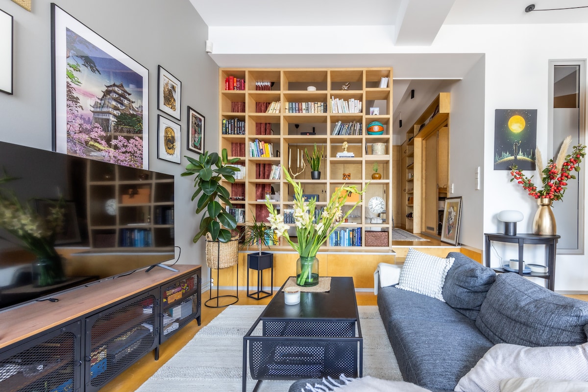 A welcoming living area features a large television on a media console, complemented by a decorative bookshelf filled with various books and artwork. A modern sofa with throw pillows faces the TV, while a vibrant plant adds a touch of greenery to the space.