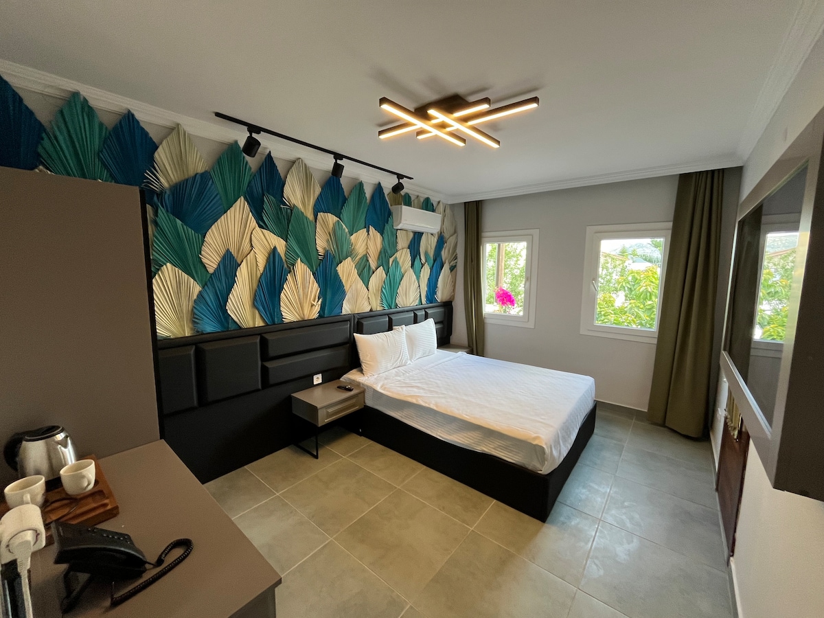 A modern room features a spacious bed against a textured wall adorned with abstract leaf designs in blue and green. Natural light enters through two windows, complemented by soft green curtains. A minimalistic desk and a flat-screen TV are positioned nearby.