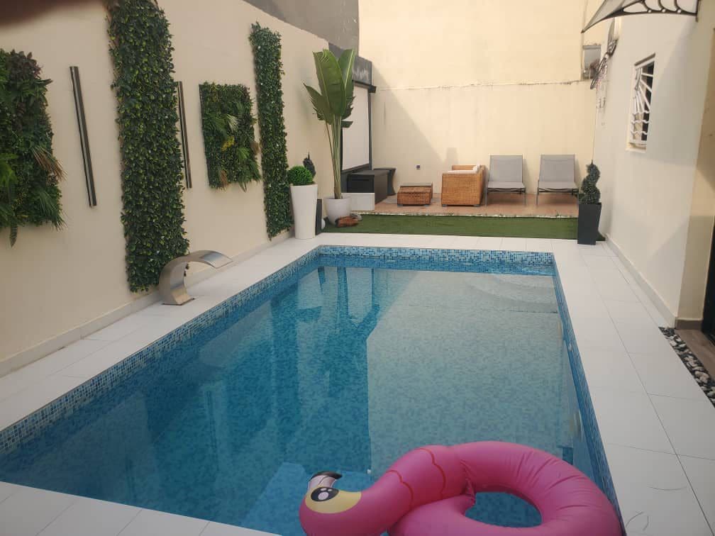 A serene outdoor area is highlighted by a clear swimming pool surrounded by white tile. Greenery adorns the walls, contributing to a relaxed ambiance. Two lounge chairs are positioned nearby, and a pink inflatable float rests in the pool, adding a playful touch to the setting.