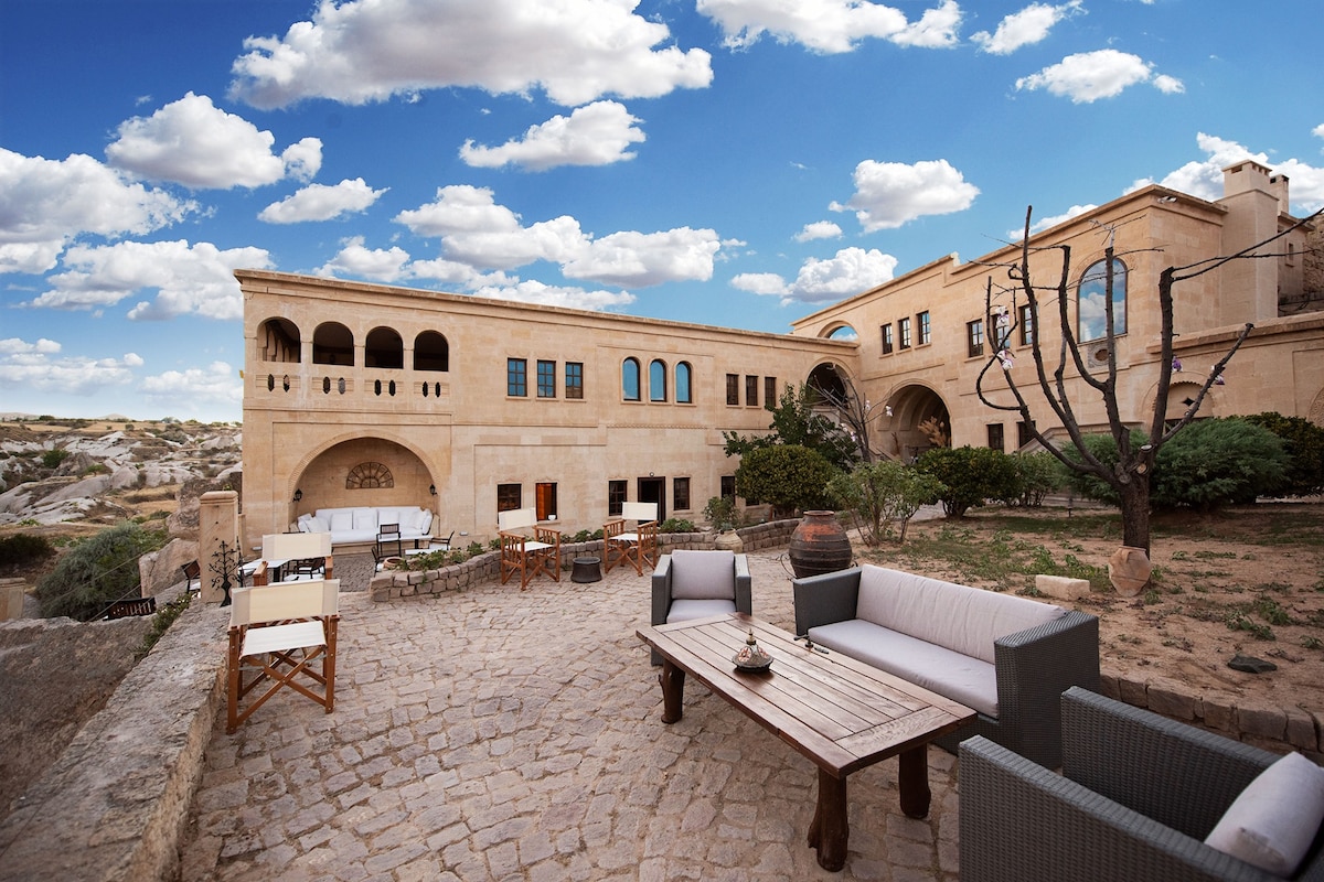 A serene courtyard is featured in the image, showcasing a variety of outdoor seating arrangements. A long wooden table is flanked by multiple chairs. Surrounding greenery adds a natural touch, while the sculptural architecture of the hotel is visible in the background under a partly cloudy sky.