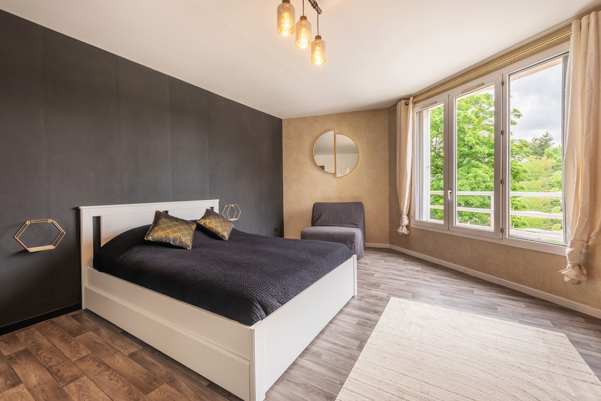 A spacious bedroom is illuminated by pendant lighting, featuring a large bed with dark bedding. A cozy chair is positioned beside the bed. Natural light fills the room through large windows, offering a view of greenery outside. A round mirror is mounted on the wall.