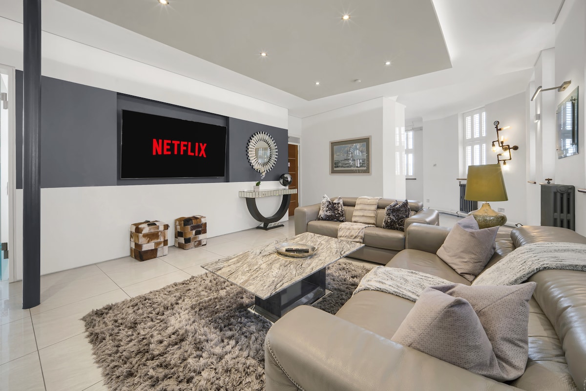 A spacious living room features a contemporary sectional sofa adorned with decorative pillows. A large, stylish coffee table sits on a plush area rug, while a smart TV is mounted on the wall, showcasing a streaming service logo. Natural light floods in through large windows.