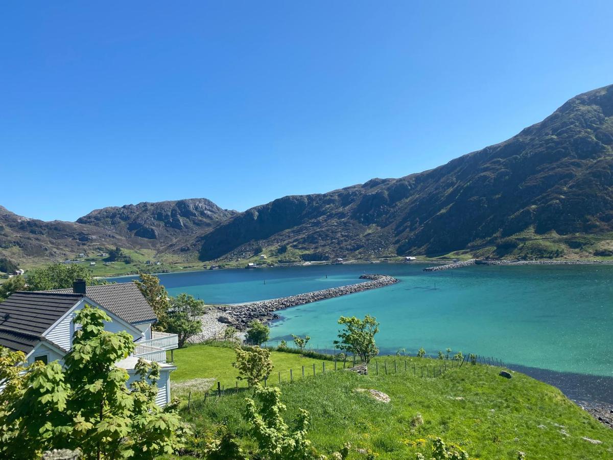 A serene landscape depicts a tranquil sea surrounded by lush green lawns and rocky mountains in the distance. Two nearby cottages are visible along the water's edge, with bright blue skies overhead enhancing the natural beauty of the rural setting.