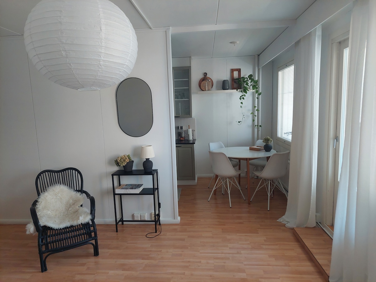 A well-lit living space features a cozy black wicker chair adorned with a soft white throw. A round dining table with four chairs is positioned nearby. A decorative plant is placed on the wall shelves, while bright curtains allow natural light to filter through.