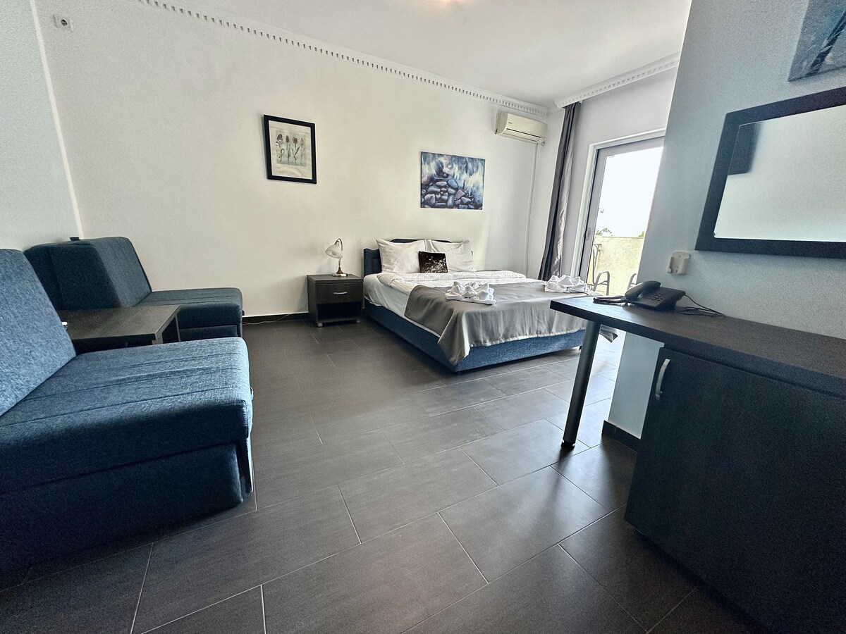 A spacious one-room accommodation features a sleeping area with a bed covered by a light blanket. A small desk and a chair are situated next to the bed, while two blue seating arrangements are placed against the wall, complemented by a large window that provides natural light.