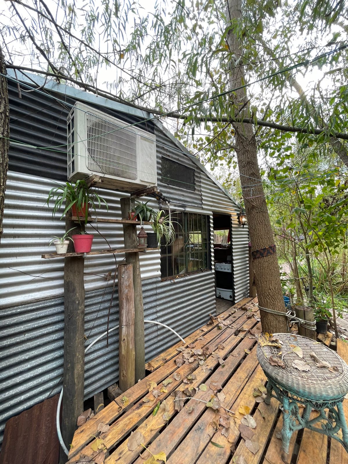 A rustic tiny house is displayed, featuring a corrugated metal exterior. A small wooden deck is visible, adorned with fallen leaves. Potted plants are positioned near the window, and an air conditioning unit is mounted on the side. Surrounding greenery contributes to a serene, natural setting.