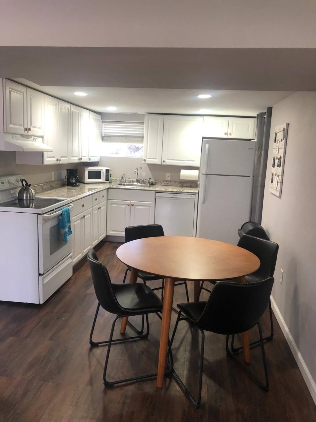 A modern kitchen with white cabinetry features essential appliances, including a stove, microwave, and refrigerator. A round wooden dining table is surrounded by four black chairs. Natural light enters through a window above the sink, enhancing the clean and functional layout.
