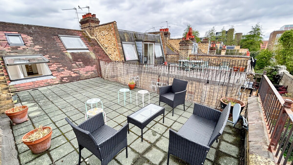 The rooftop terrace is spacious, featuring outdoor furniture including a glass-top table and several chairs. Potted plants add greenery to the area, while views of nearby buildings and the city skyline can be observed. The terrace is enclosed by a low fence.