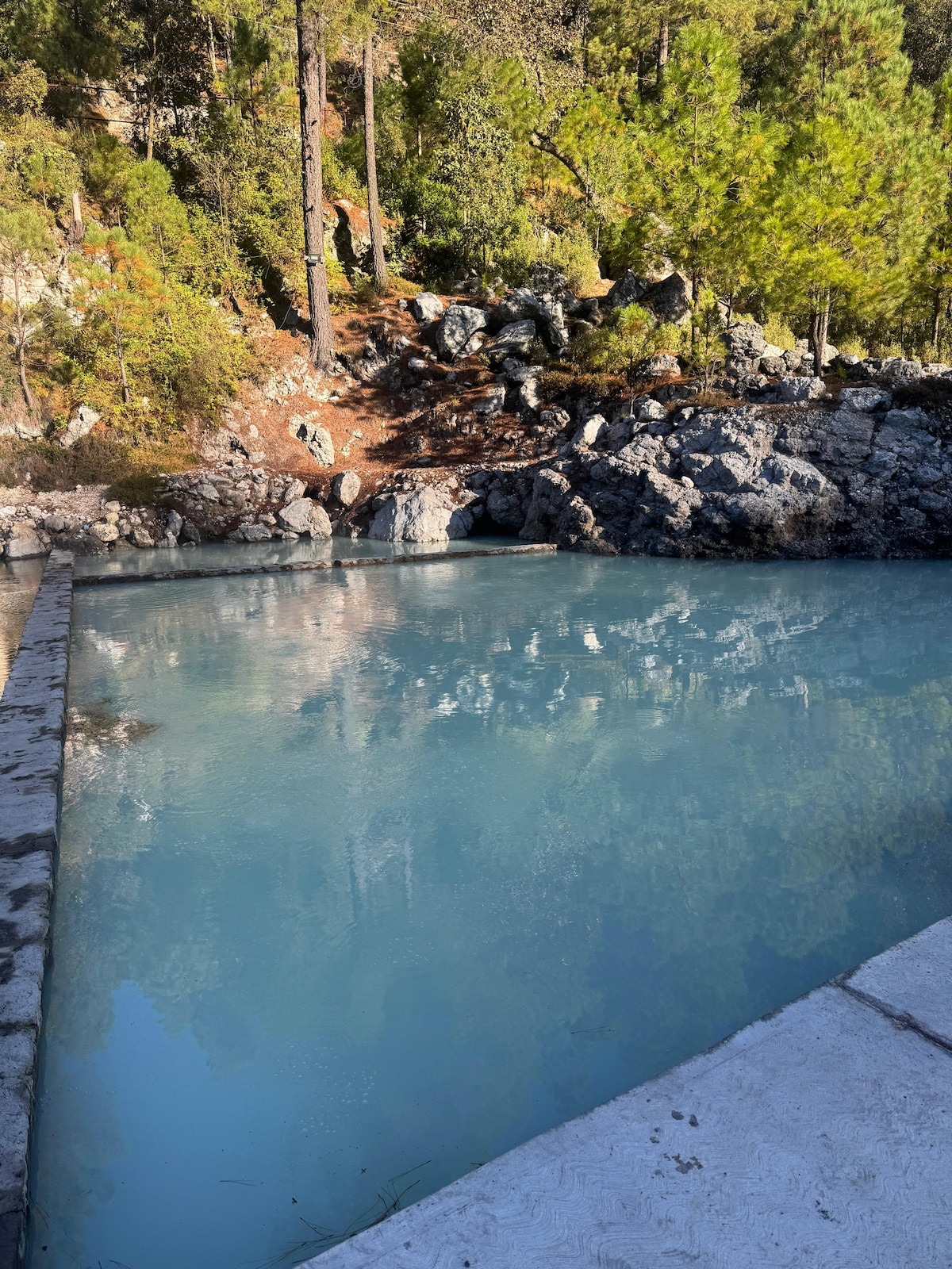 A serene swimming area is surrounded by rocky terrain and lush greenery. The water appears calm and reflects the surrounding trees, creating a peaceful environment. The stone edge of the pool is visible, contrasting with the natural landscape.