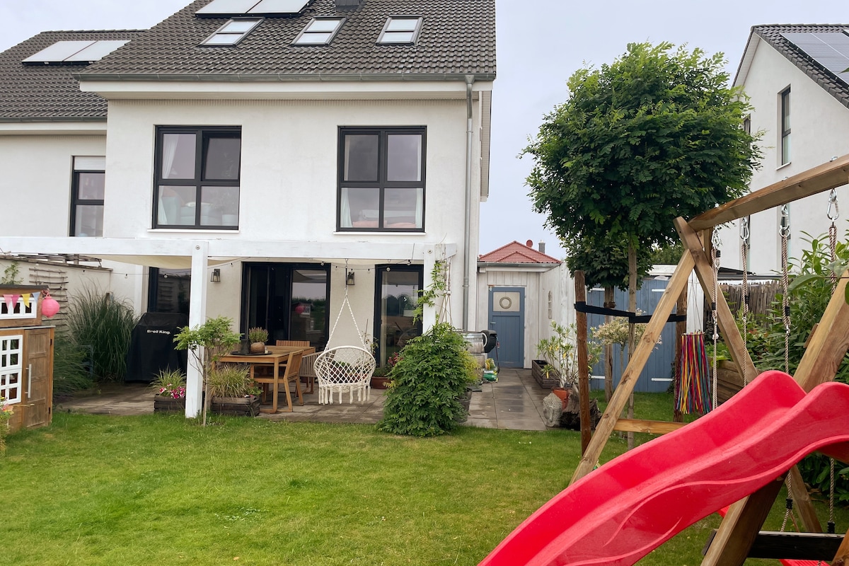 The exterior of the home showcases a two-story house with large windows. A spacious garden features a playful slide, a swing set, and a seating area with a table and chairs under a covered patio, surrounded by greenery and plants.