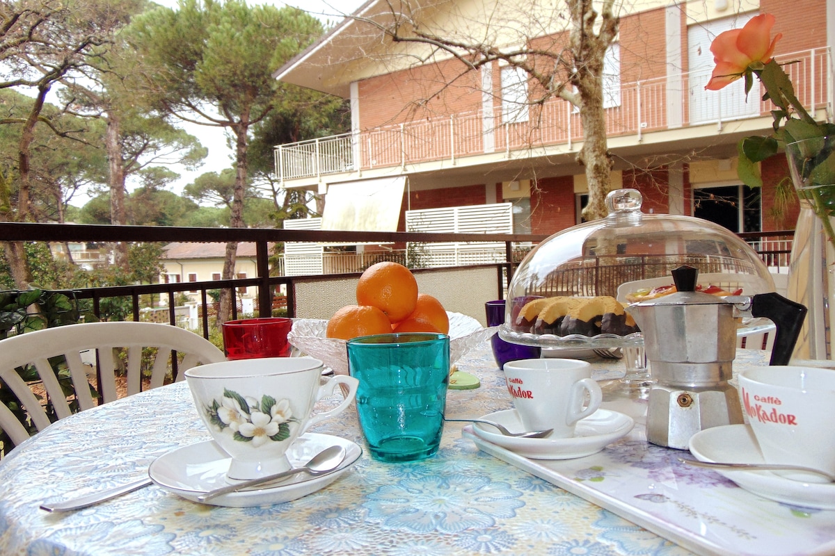 A table is set on a balcony overlooking a garden, adorned with a floral tablecloth. A glass pitcher, coffee pot, and assorted pastries are present alongside fruits. The scene is framed by greenery and features a view of the surrounding buildings.