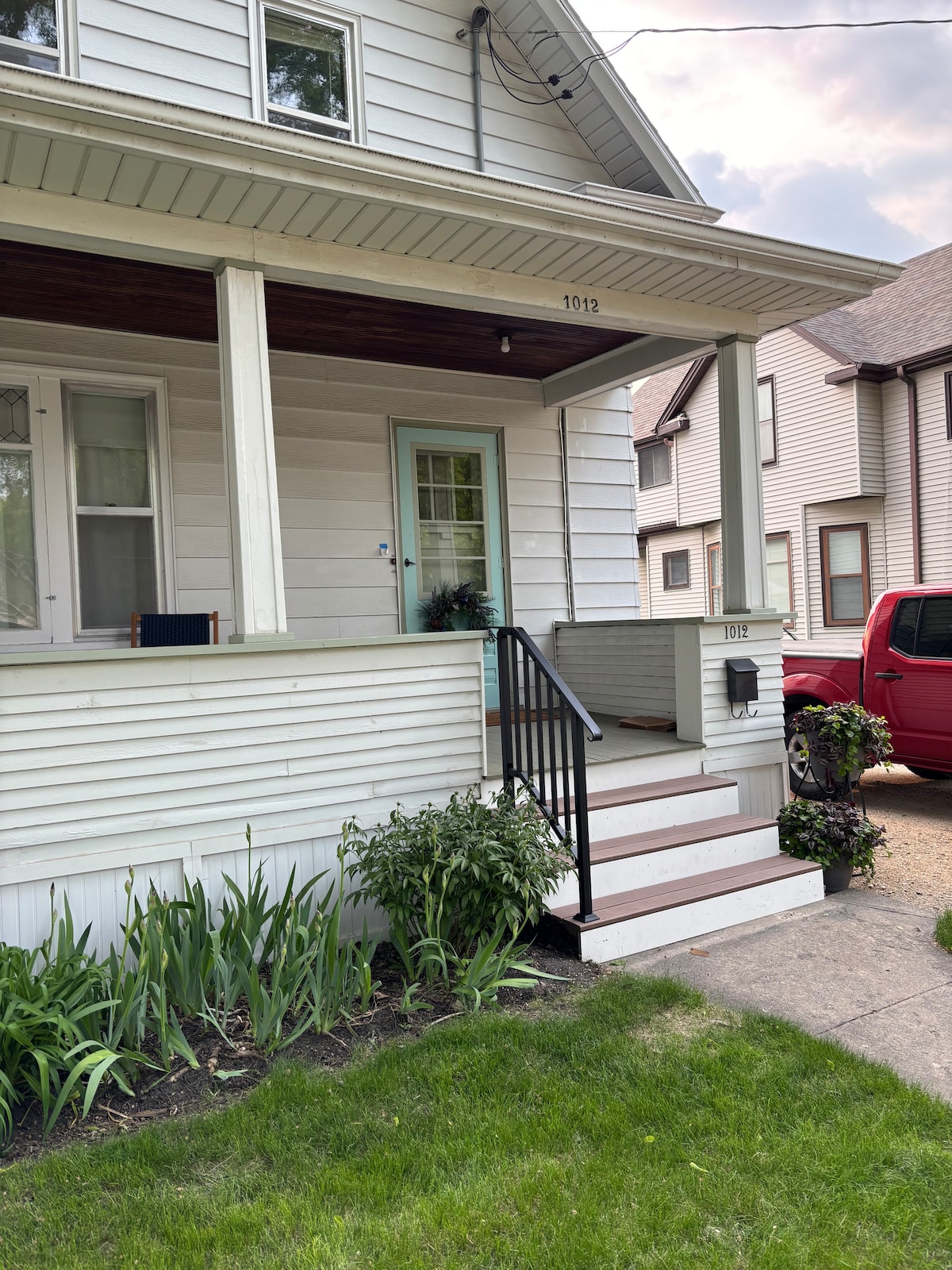 The entrance of a two-story house is showcased, featuring a covered porch with steps leading up to the front door. Flanking the entrance are modest flowerbeds with greenery. A seating area is visible on the porch, and a pathway is seen leading to the driveway.