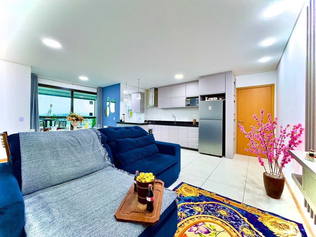 A well-appointed living area features a large blue sofa with a light throw, accompanied by a decorative tray with snacks and beverages. A modern kitchen with sleek cabinetry is visible in the background, complemented by a potted plant with pink blossoms and a vibrant area rug.