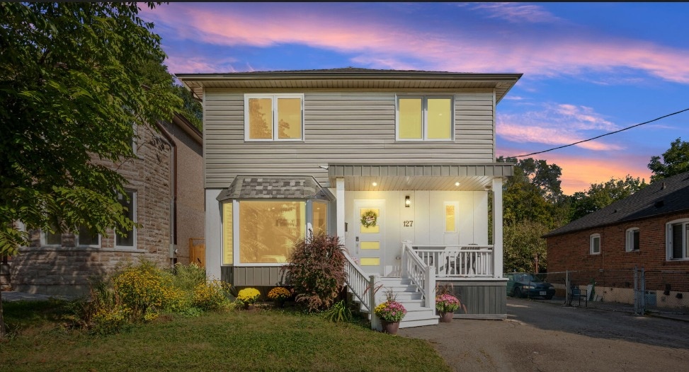 A spacious two-story home is depicted, featuring a covered porch adorned with potted plants. Large windows allow natural light to enter, complementing the exterior's neutral tones. A lush front yard with vibrant flowers adds to the welcoming presence of the property, set against a colorful evening sky.