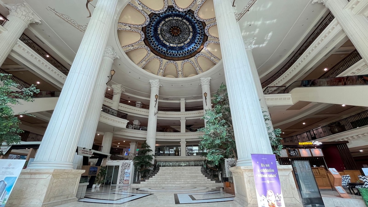 An indoor lobby is showcased, featuring large white columns and a high ceiling adorned with a decorative circular design. Elegant stairs lead to upper levels, flanked by lush green trees. The area is open and well-lit, creating a spacious and welcoming environment.