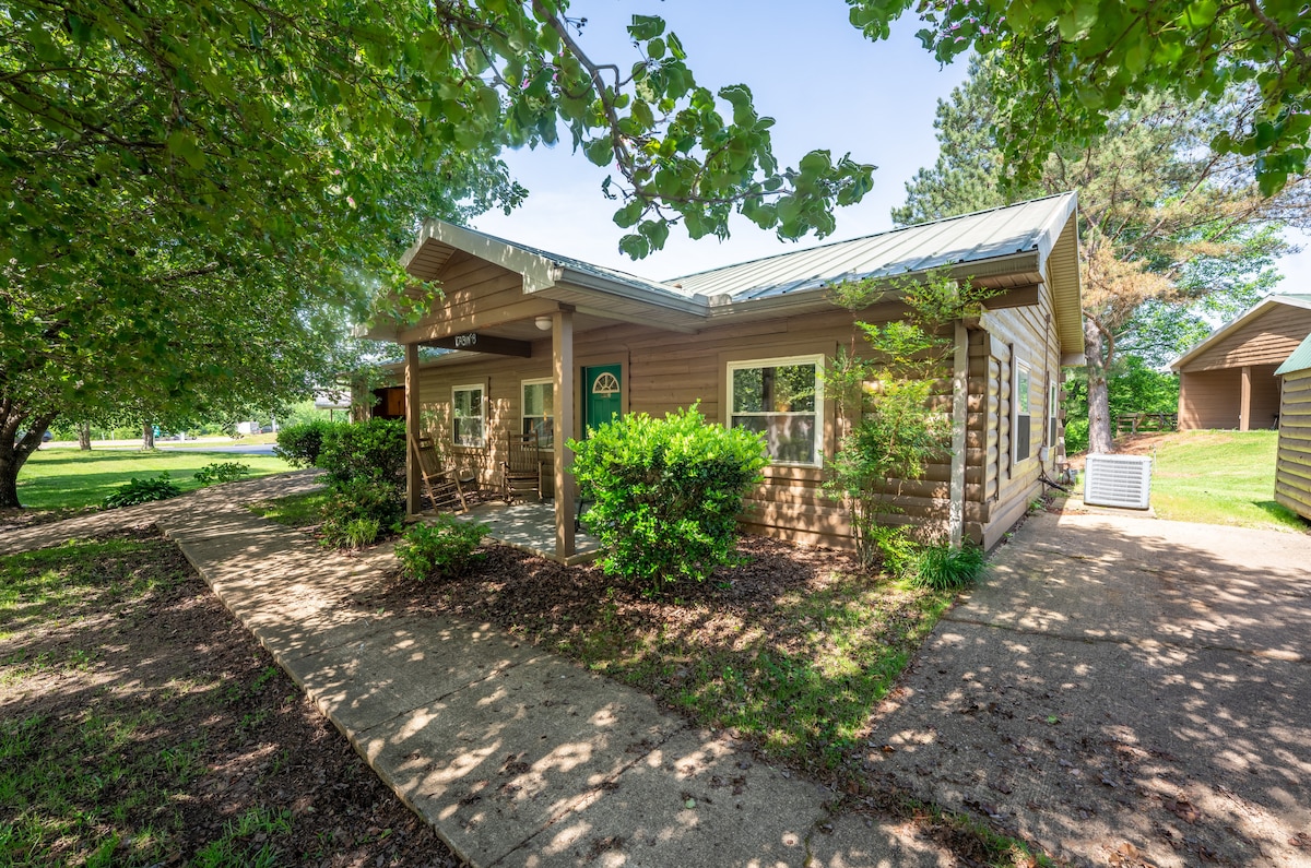A charming cabin exterior is framed by lush green trees and shrubs, showcasing a shaded porch with seating. The light-colored wooden siding and metal roof complement the inviting entranceway, which features a decorative door. A clear pathway leads to the entrance, surrounded by manicured landscaping.