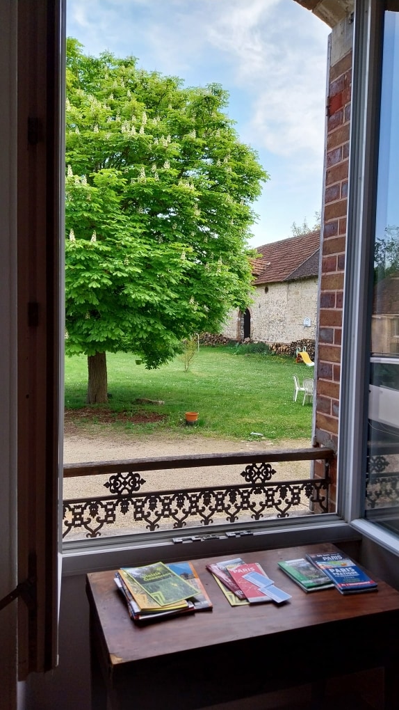 A window opens to a view of a lush green tree in the garden, with a well-manicured lawn visible. A selection of books rests on a wooden table inside, complementing the serene outdoor setting.