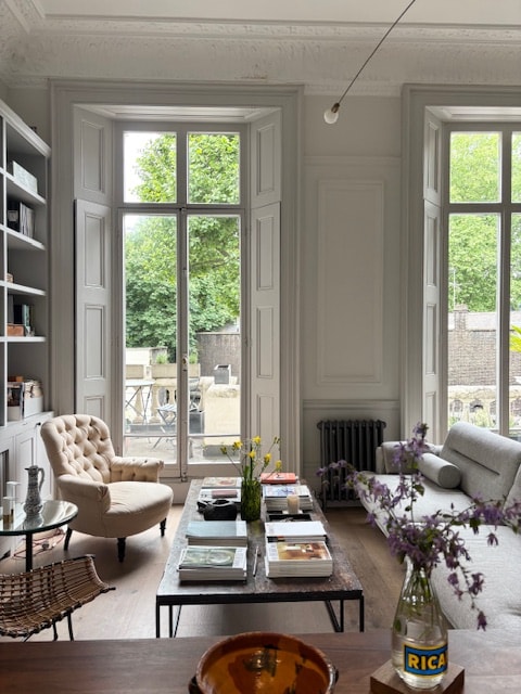A spacious sitting area includes a light-colored armchair beside a wooden coffee table, which is topped with books and a decorative bowl. Large windows with wooden shutters provide ample natural light and offer views of greenery outside. A neutral sofa is positioned nearby.