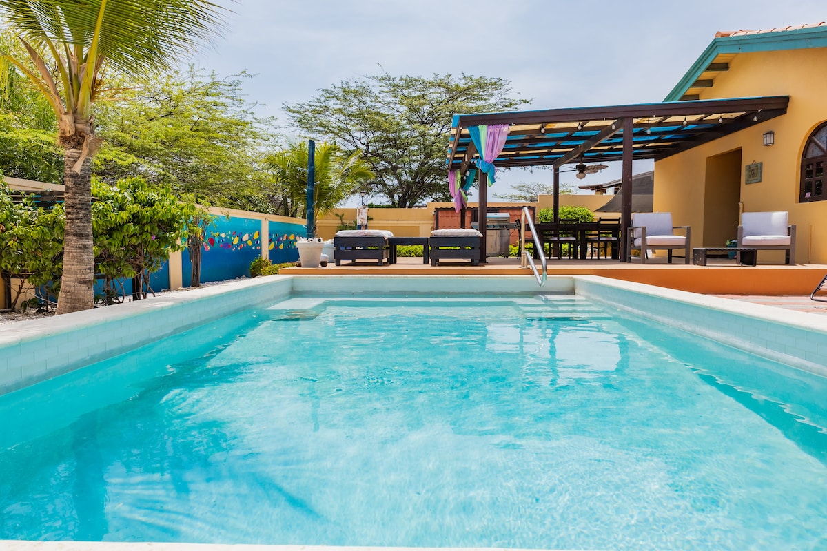 A private outdoor patio features a glistening pool with inviting aqua water. Comfortable seating areas are visible alongside a dining space under a covered structure, surrounded by lush greenery and vibrant colors, creating a serene retreat for relaxation.
