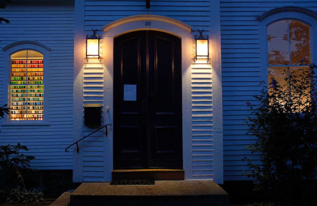 The entrance of the building is illuminated by two wall-mounted lanterns, casting a soft glow. The large double doors are a dark shade, while colorful stained glass can be seen through the arched windows on either side, highlighting the architectural details.