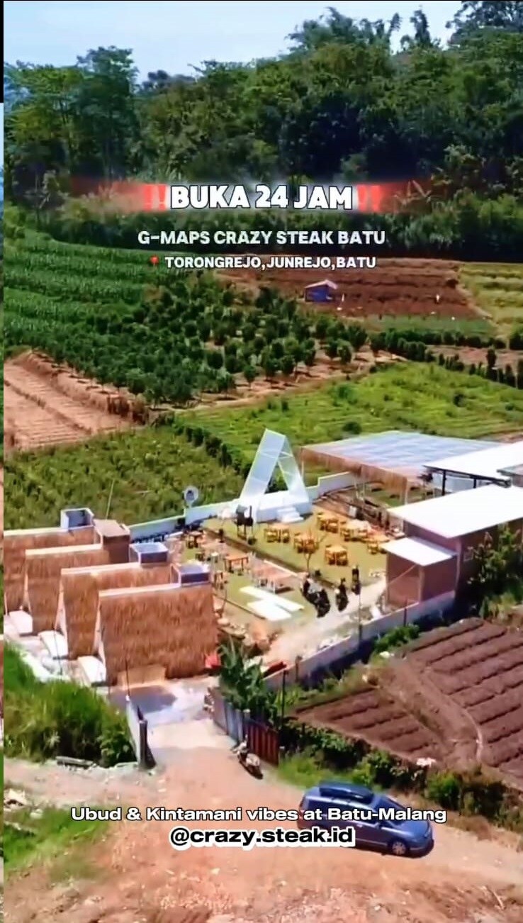 A scenic outdoor dining area surrounded by lush green fields. Thatched-roof structures are positioned alongside wooden tables and chairs, offering a communal atmosphere. A clear sky is visible above, enhancing the natural beauty of the setting.
