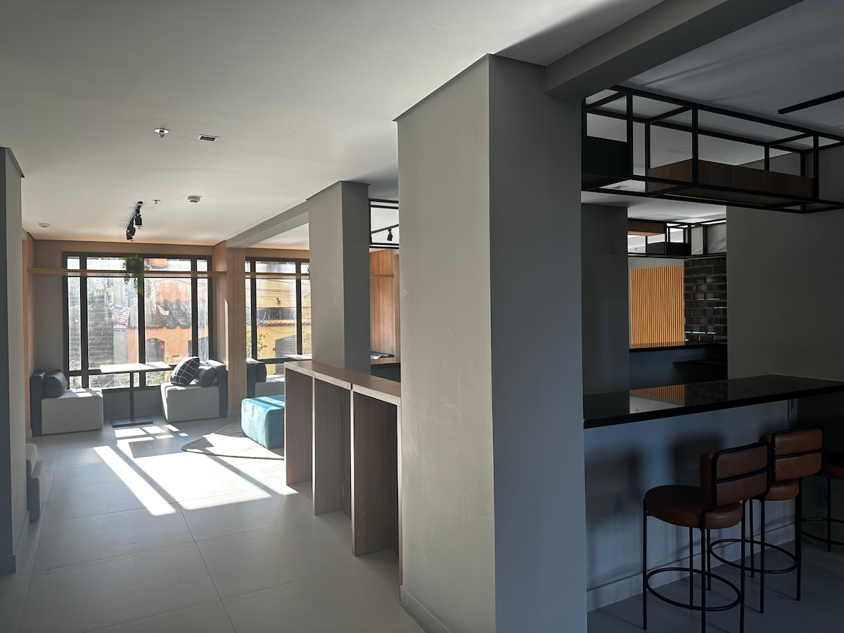 An open layout showcases a modern studio space with large windows providing natural light. A kitchen bar with high stools is visible, alongside a cozy seating area with contemporary furniture. The versatile design creates an inviting atmosphere, suitable for relaxation or casual dining.