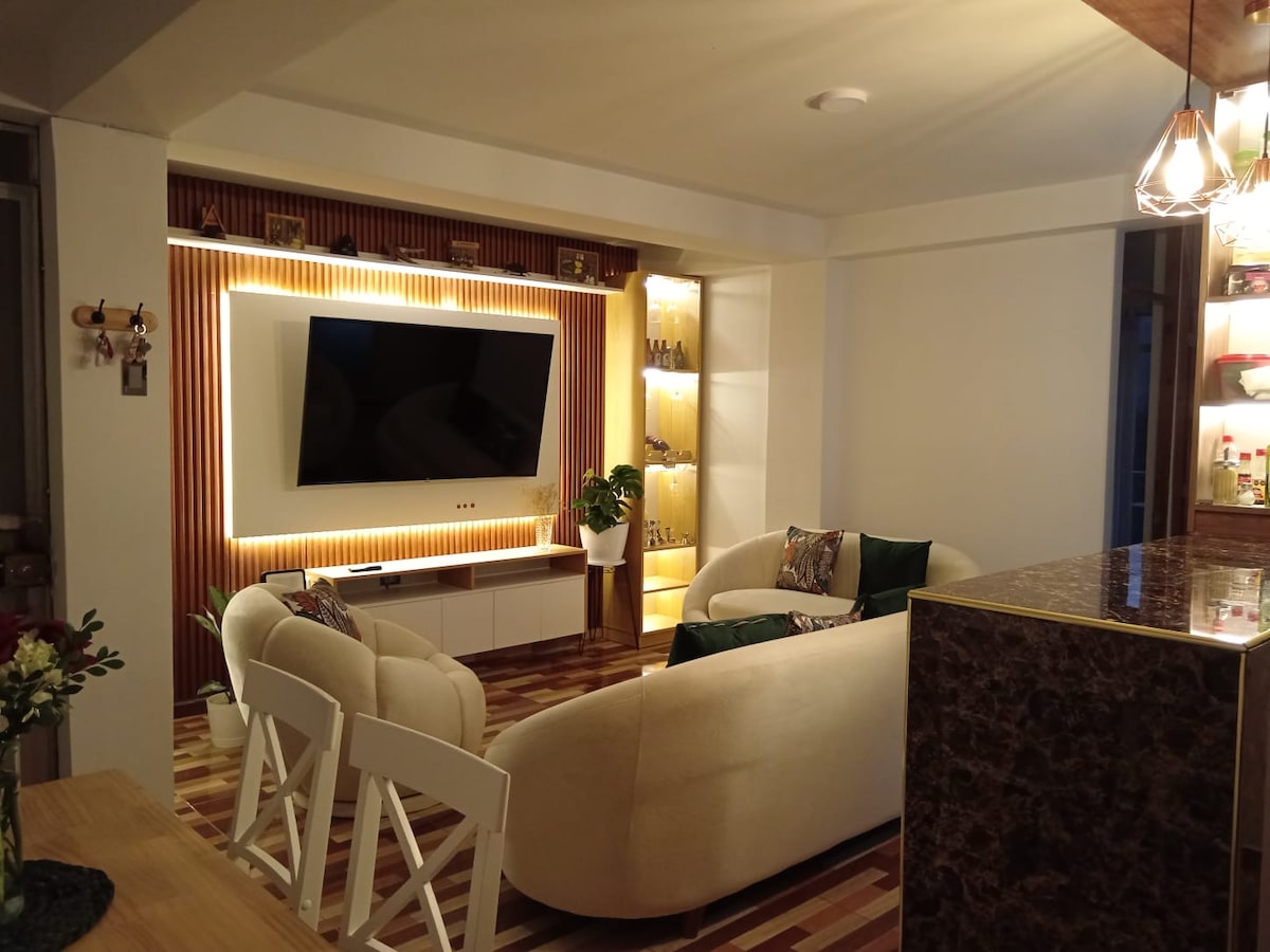 A modern living area features a large television mounted on a wooden accent wall, illuminated by soft lighting. Two curved sofas in a light tone are arranged around a small table, complemented by decorative shelving that displays items. Natural light enters through adjacent windows.