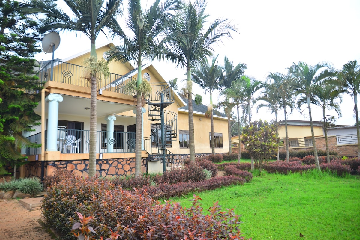 The exterior of a spacious house is surrounded by lush greenery and well-maintained gardens, featuring palm trees and shrubs. A spiral staircase leads to a second level, while a patio area is visible, suggesting opportunities for relaxation and outdoor dining.