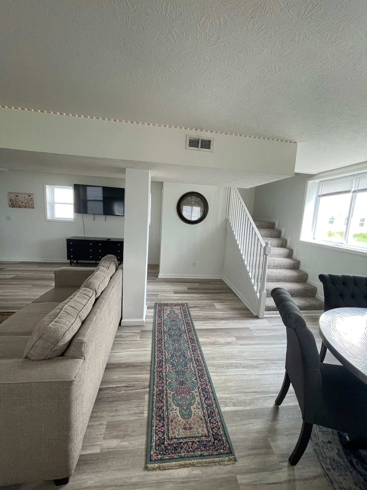 A spacious living area is furnished with a comfortable sofa facing a wall-mounted television. A decorative rug runs along the floor, leading toward a circular dining table beside a large window. A staircase can be seen in the background, illuminated by natural light.