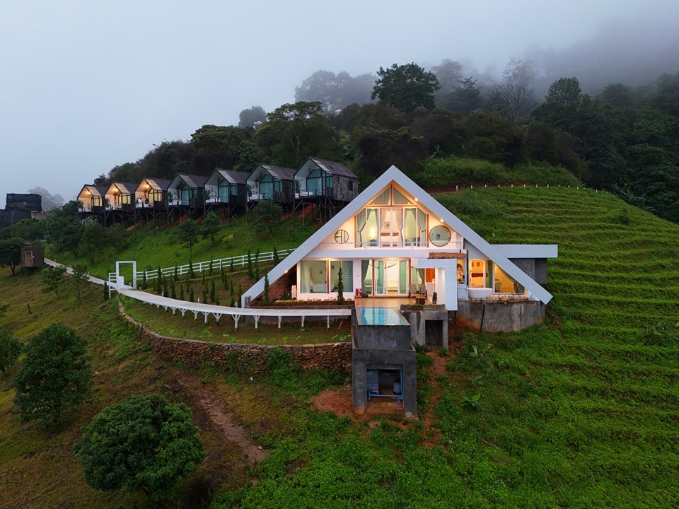 A modern villa is positioned on a hillside, featuring a distinct triangular roof and large windows. Surrounding the villa, lush greenery is visible, along with several separate accommodations in the background. The scene is enveloped in soft morning mist.