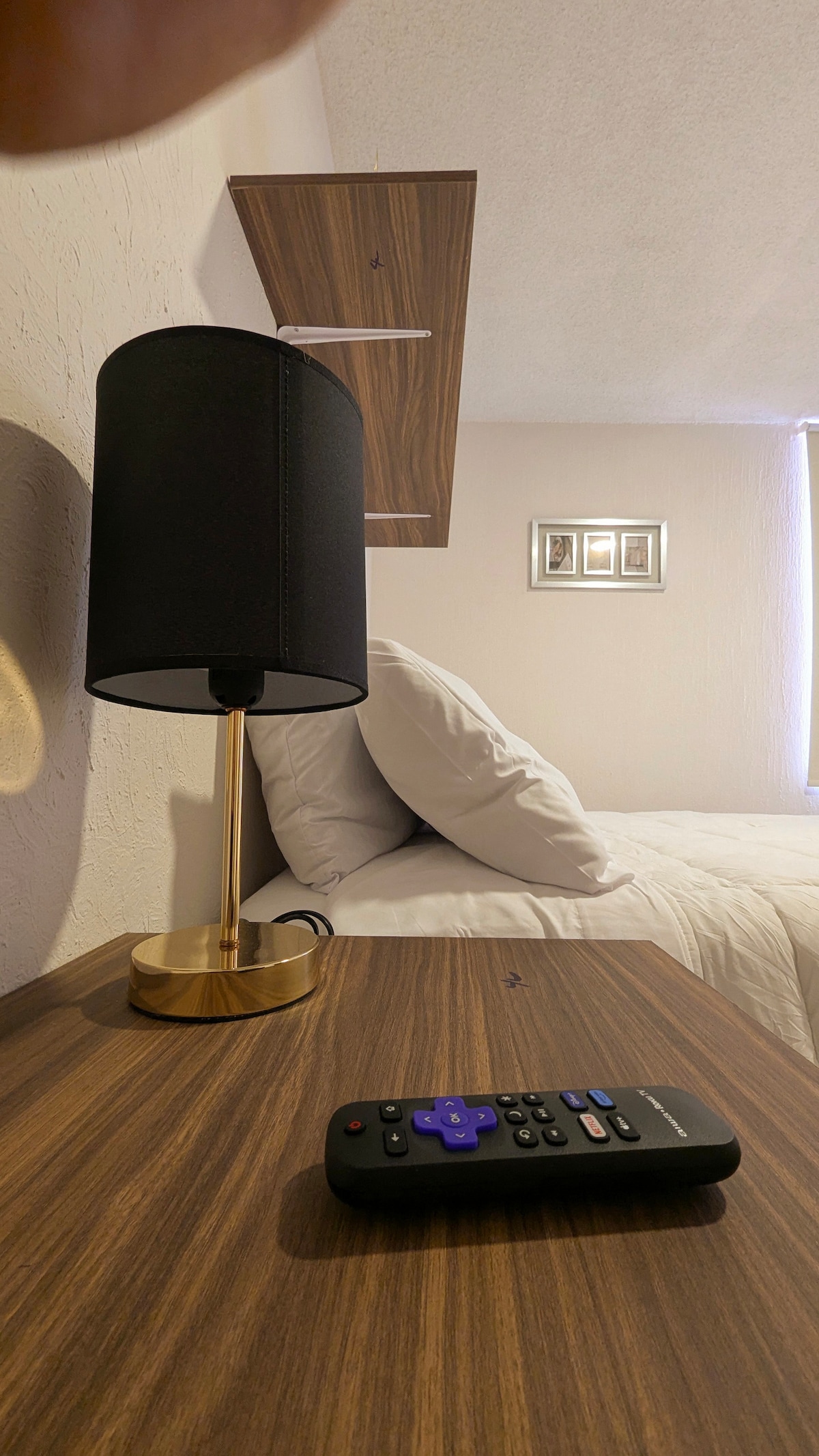 A wooden bedside table holds a sleek, gold lamp with a black shade next to a spacious bed adorned with white pillows and bedding. A remote control is placed on the table, and soft natural light enters through a nearby window.