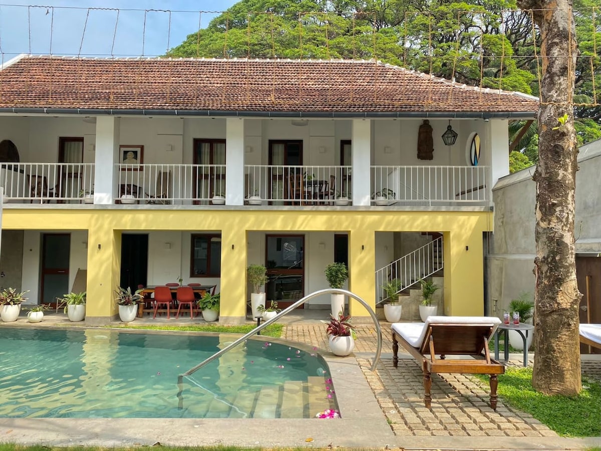 The exterior of the villa showcases two levels with a charming yellow façade. A private pool is surrounded by a stone patio, featuring a sunbed and a small wooden table. Lush greenery frames the scene, enhancing the serene atmosphere.