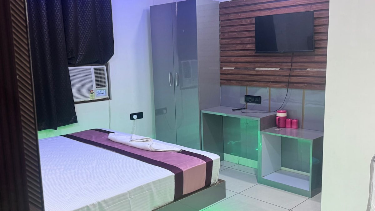A bedroom is presented with a spacious bed covered in light bedding. A wardrobe and a television are mounted on the wall, with a console underneath. An air conditioning unit is positioned by the window, and soft lighting adds a calm ambiance.