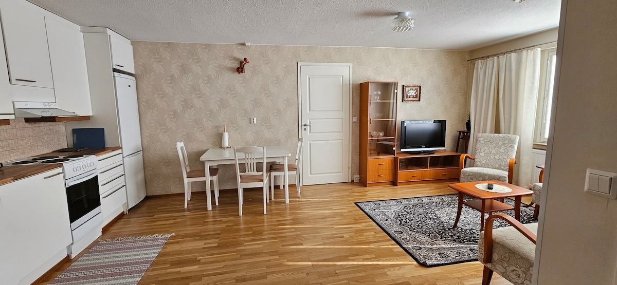 An open living area displays a blend of modern and classic furnishings. A dining table with white chairs is positioned next to a wooden coffee table. A television is set upon a wooden console. Natural light enters through large windows, illuminating the polished wooden floor.
