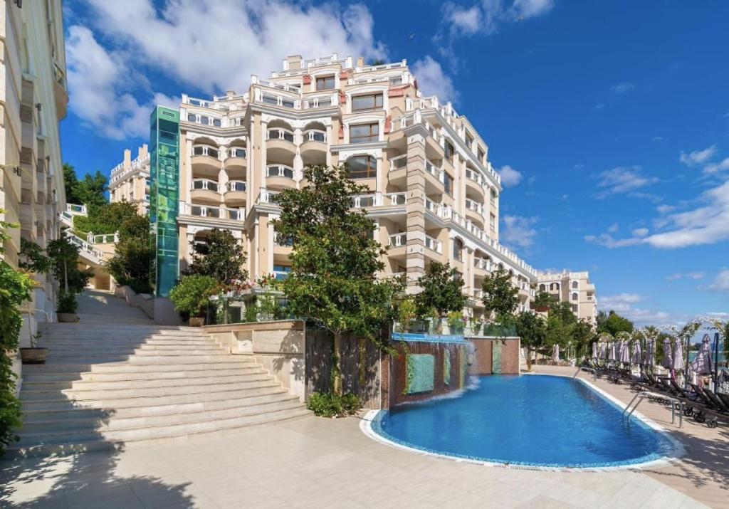 A large resort complex is shown with a pool in the foreground, featuring clear blue water. Steps lead from the poolside up to the main building, which has multiple stories and a mixture of greenery and terrace areas. The sky is bright with scattered clouds.