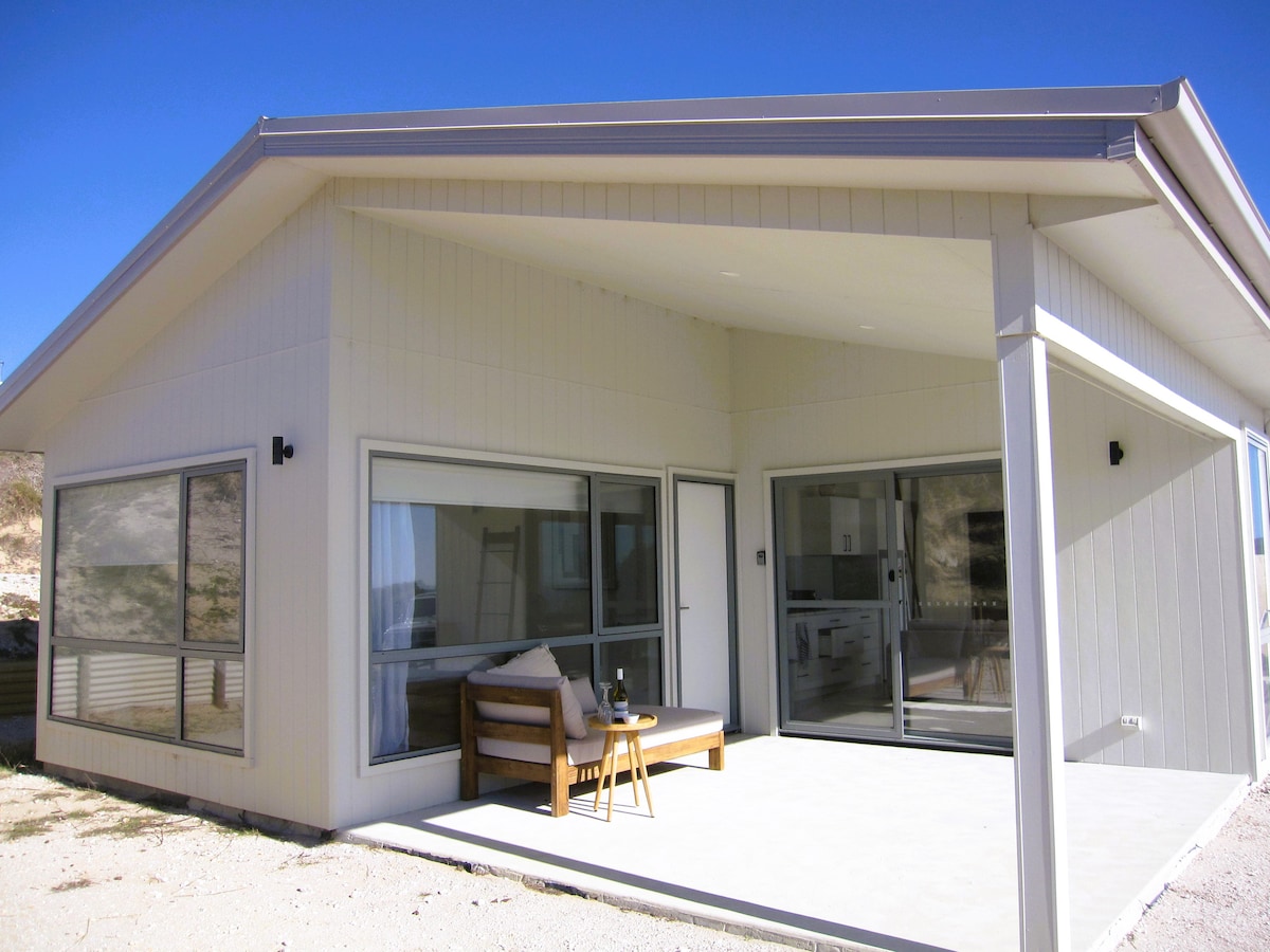 A modern cabin exterior features a spacious porch with cozy seating. Large windows provide ample natural light and an inviting view of the surroundings. The clean lines of the architecture contribute to a relaxed atmosphere, with a subtle connection to the outdoors.