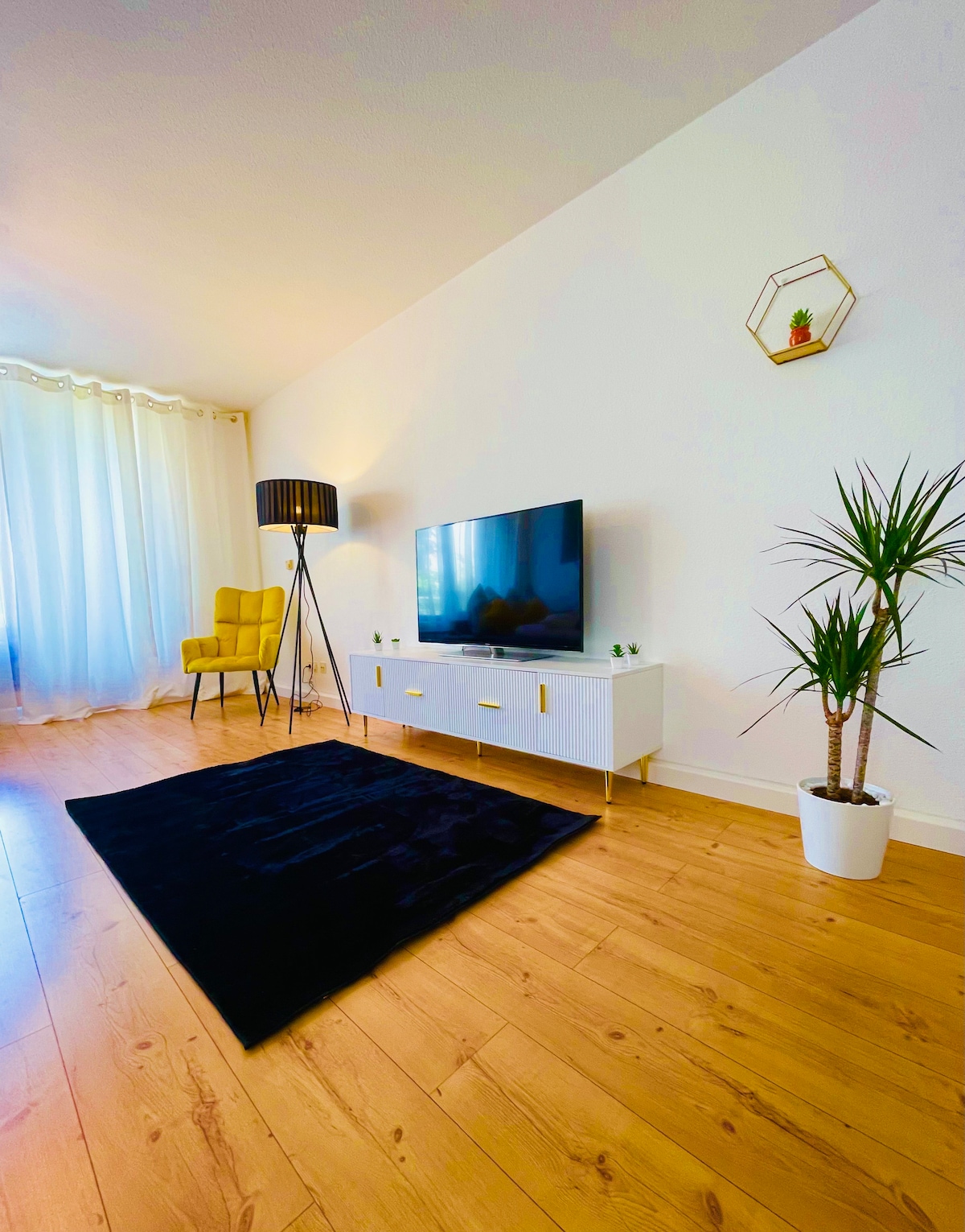 A bright living area features a comfortable armchair and a modern TV stand with a flat-screen television. A tall lamp stands beside the chair, contributing to the cozy atmosphere. Large windows allow for natural light to filter in, with light curtains draped elegantly.
