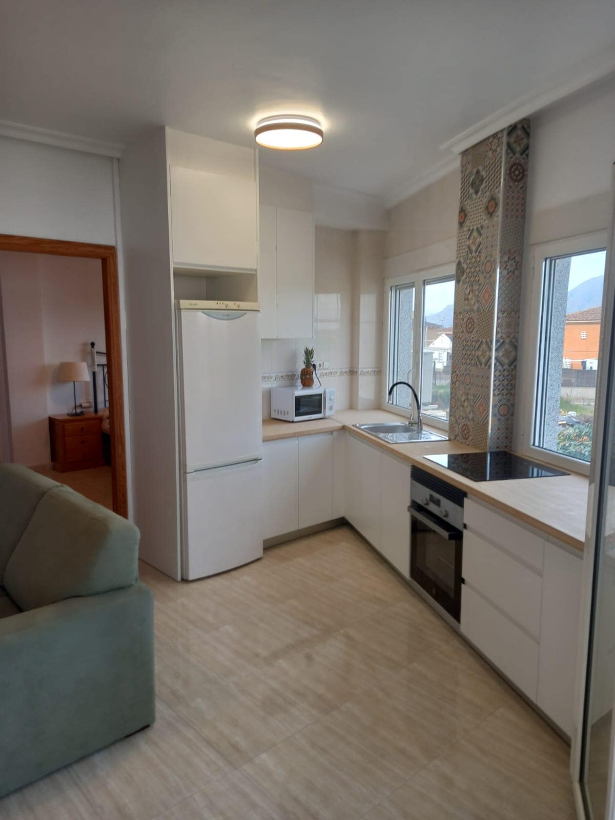 A modern kitchen is displayed, featuring sleek white cabinetry and a stainless-steel microwave. Natural light enters through multiple windows, illuminating the spacious counter with a sink and a built-in oven. Decorative tiles add a subtle touch of design to the room.