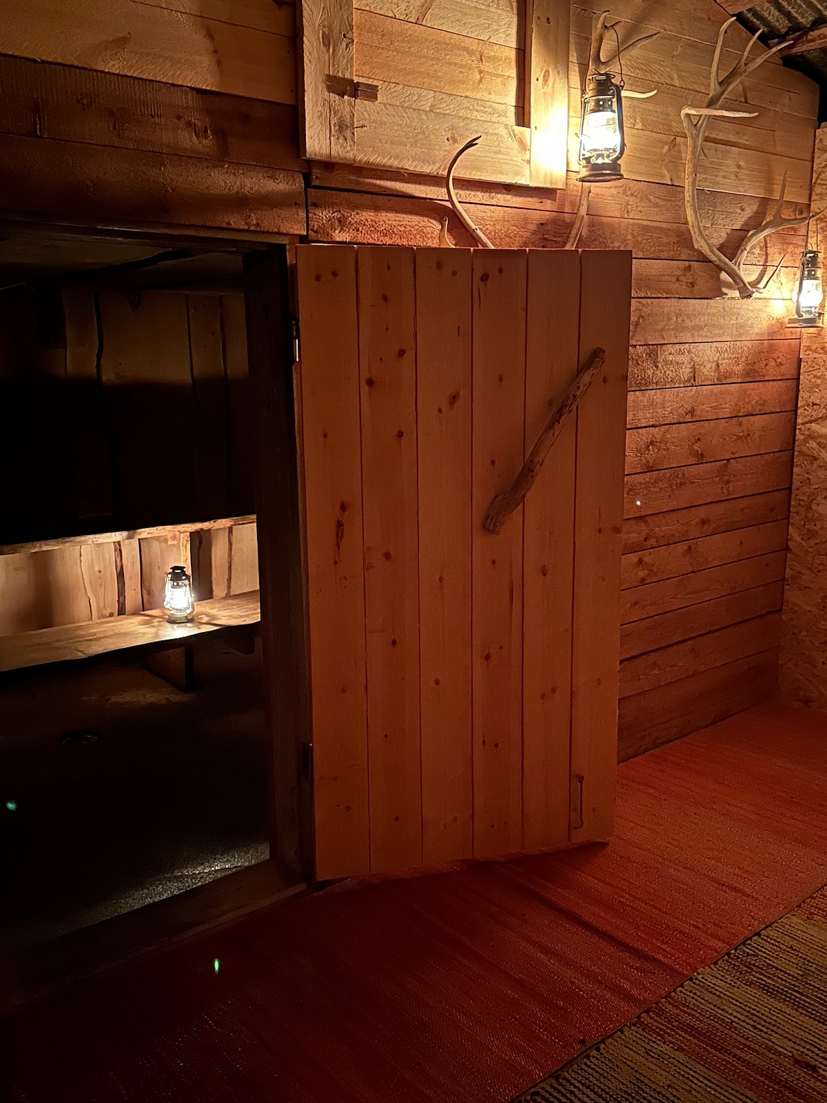 An entrance to a wooden sauna is visible, featuring a rustic door crafted from planks and adorned with a simple wooden handle. Lanterns hang on the walls, casting a warm glow over the interior space, which is warmly lit to create a cozy atmosphere.