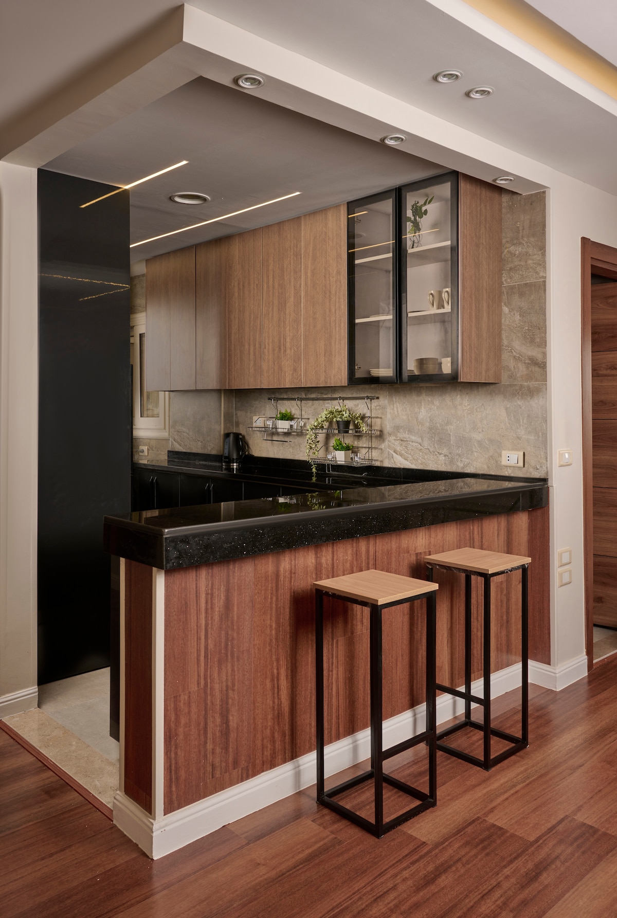 A modern kitchen area features sleek cabinetry in warm wood tones, complemented by a black countertop. At the bar, two high stools provide additional seating. Natural light is introduced through a nearby window, enhancing the overall design with a blend of functionality and style.