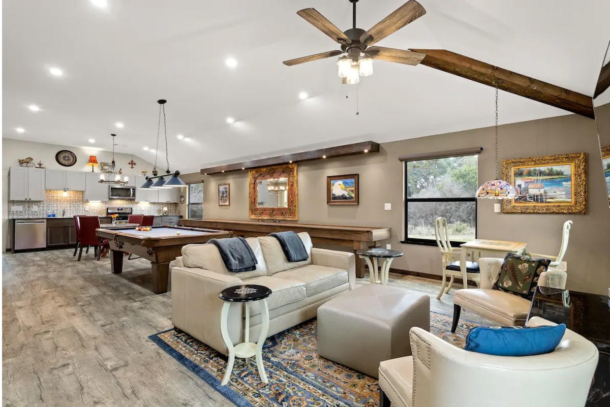 A spacious living area is highlighted by a large pool table and comfortable seating. Natural light fills the room through expansive windows, illuminating the warm, neutral tones of the decor. Unique artwork and light fixtures add character to the inviting atmosphere.