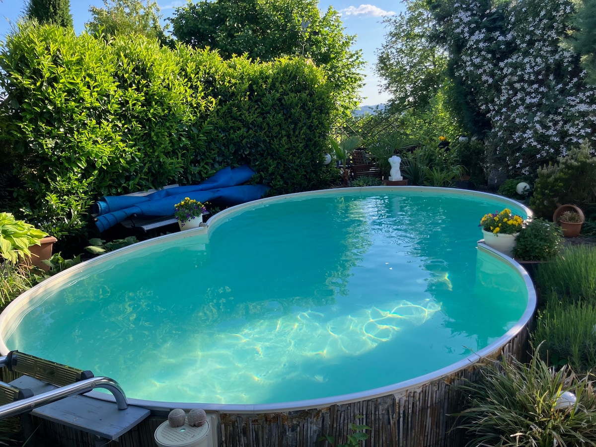 A tranquil swimming pool is set within a lush garden, surrounded by various greenery and flowering plants. The pool's water glistens in the sunlight, reflecting the clear blue sky. Lounge areas with seating can be discerned in the background, inviting relaxation.