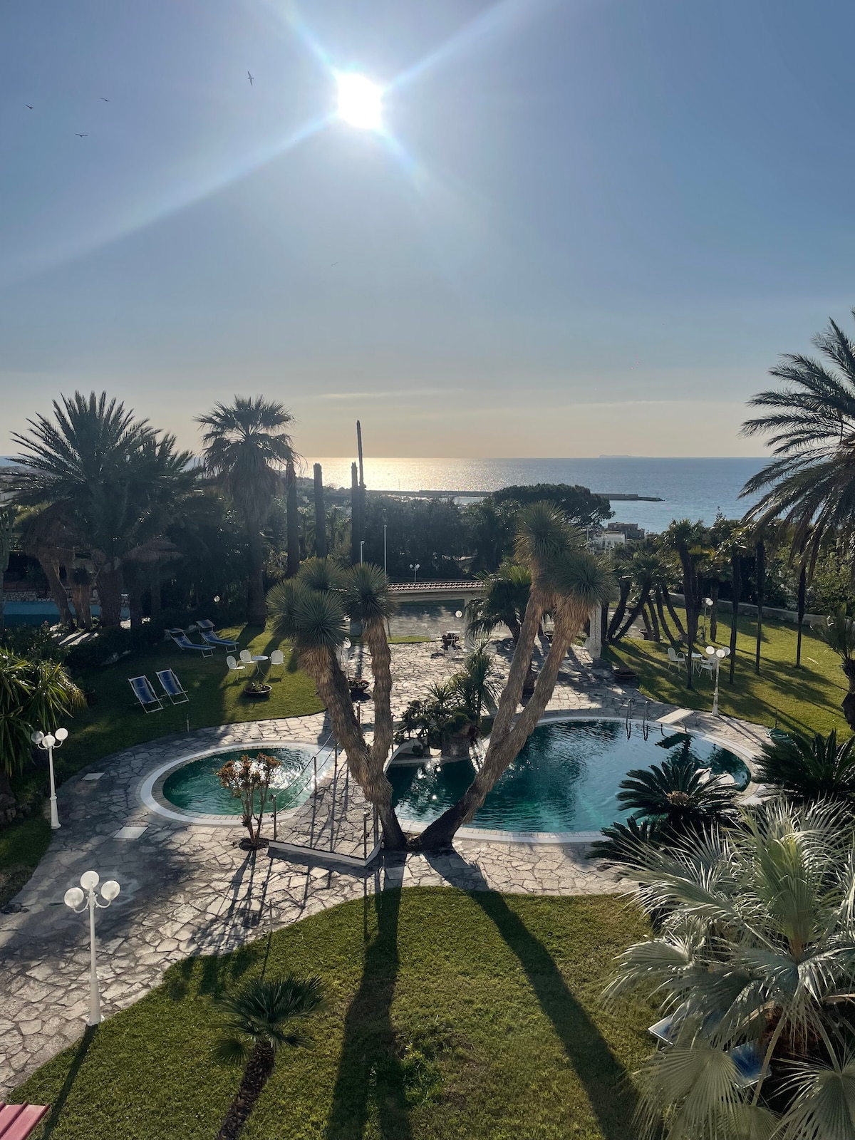 A panoramic view features a sparkling pool surrounded by palm trees and well-maintained landscaping. The sparkling sea is visible in the background under a bright sun. Outdoor lounge chairs and light fixtures enhance the serene atmosphere, inviting relaxation in this elegantly arranged space.
