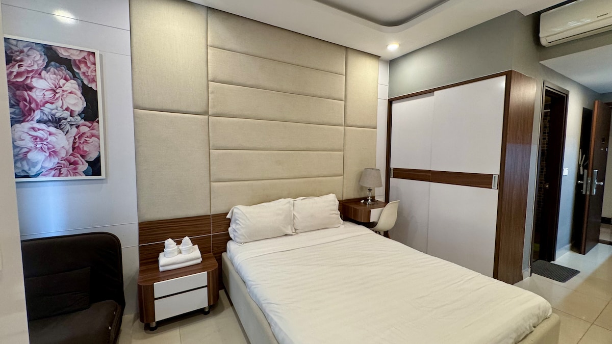 A modern sleeping area features a spacious bed with a neutral-colored headboard. A bedside table with a lamp accompanies the bed, while a sleek storage unit is integrated into the room's design. Floral artwork adorns the wall, adding a subtle touch of color.